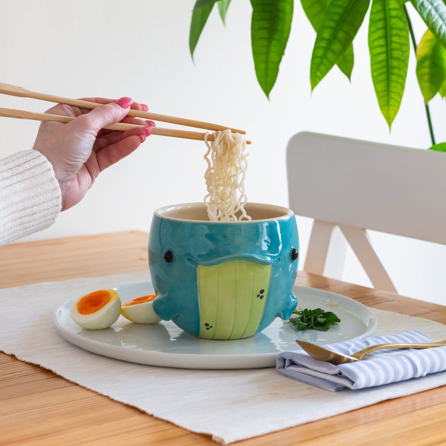 Person using chopsticks to eat from a blue whale ceramic bowl on a table with a plant in the background.