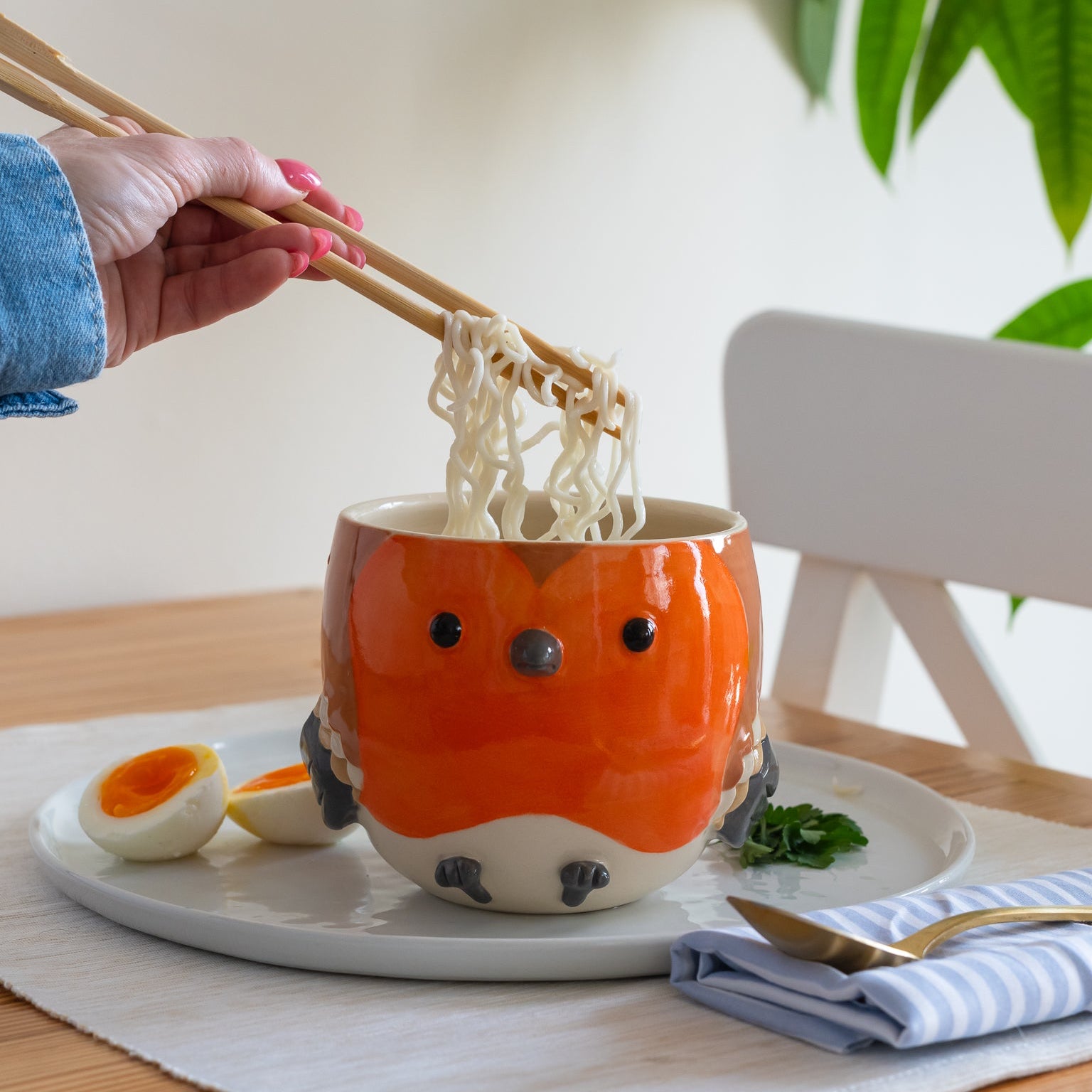Person using chopsticks to eat noodles from a sculpted robin bowl on a table.