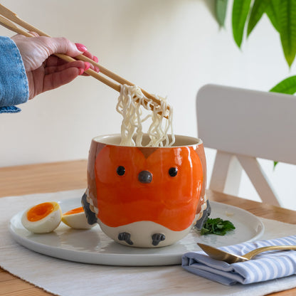 Person using chopsticks to eat noodles from a sculpted robin bowl on a table.