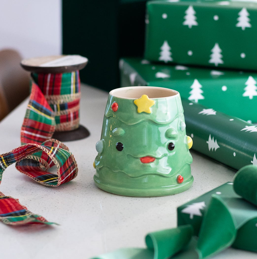 Green ceramic cup with Christmas tree design, green gift wrap, and ribbon on a white surface.
