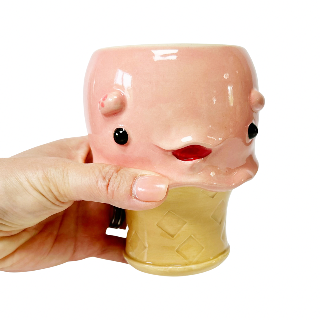A hand holding a pink ceramic novelty mug shaped like an ice cream cone, against a plain white background.